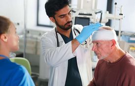 Schädel-Hirn-Trauma: Symptome & Behandlung Arzt untersucht älteren Patienten mit Kopfverband, weibliche Pflegekraft steht daneben