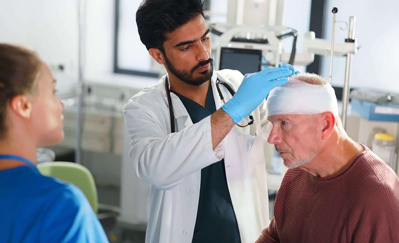 Schädel-Hirn-Trauma: Symptome & Behandlung Arzt untersucht älteren Patienten mit Kopfverband, weibliche Pflegekraft steht daneben